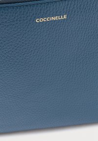 Mala de couro azul com superfície texturizada. Logotipo dourado 'Coccinelle' no topo. Forma retangular com linhas limpas.