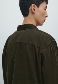 Camicia in corduroy verde scuro con colletto classico, dettaglio con bottoni sul retro e vestibilità rilassata, caratterizzata da un tessuto testurizzato e maniche lunghe.