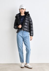Giacca a vento nera con cappuccio foderato in pelliccia, chiusura con zip, abbinata a jeans azzurri chiari e sneaker argentate. Cappellino bianco aggiunge un accento informale.