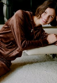 Hoodie zippé en velours marron avec une poche avant, coupe décontractée, porté sur un bas assorti. Texture douce, coutures minimales et design à capuche.