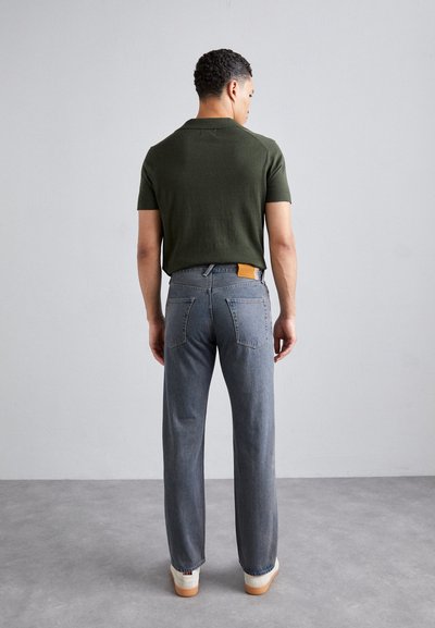 Camisa verde de manga corta combinada con unos pantalones vaqueros de color azul claro. Los vaqueros tienen dos bolsillos traseros y una etiqueta de cuero marrón en la cintura. Zapatos blancos visibles.