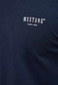 Tumesinine puuvillane T-särk, millel on valge "MUSTANG" kiri ja allpool "SINCE 1932". Pehme tekstuur ja klassikaline ümmargune kaelus.