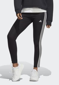 Sorte leggings med tre hvide striber langs siderne, der har en tætsiddende pasform og er parret med hvide sportsko.