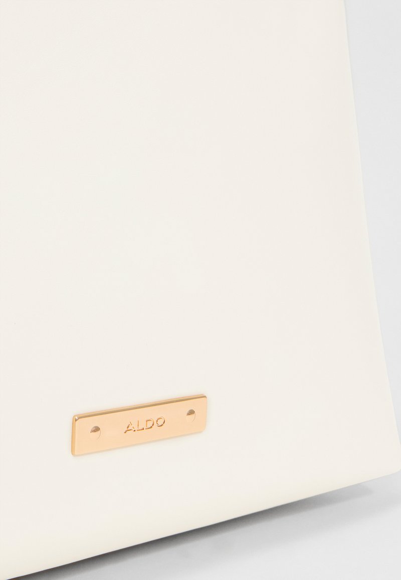 Surface blanche avec une petite plaque rectangulaire en métal doré gravée du nom de la marque "ALDO."