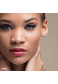 Porträtt av en person med slät hy, gröna ögon och mörk eyeliner. Läpparna är röda med en glansig finish. Händerna håller försiktigt om nacken.