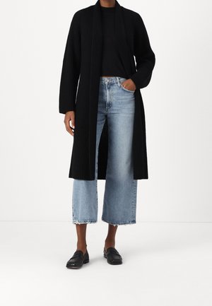 Person wearing a long black ribbed cardigan, black top, light blue wide-leg cropped jeans, and black slip-on shoes on white background.