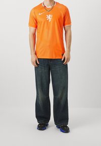 Chemise de sport Nike orange avec un emblème de lion, manches courtes et texture ponctuée, associée à un jean bleu ample et des baskets noires.