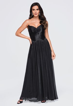 Vrouw met lang donker haar, zilveren oorbellen en een zwarte strapless geplooide avondjurk, staand met één hand op haar heup.