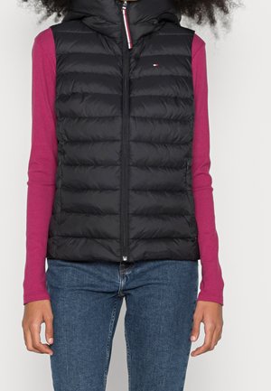 Femme portant un gilet matelassé noir à fermeture éclair sur un haut à manches longues magenta et un jean bleu, debout devant un fond blanc.