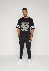 Μαύρη κοντομάνικη μπλούζα με εκτύπωση "CHAMPION" και "92" σε ασημί, σε συνδυασμό με μαύρες φόρμες και λευκά αθλητικά παπούτσια.