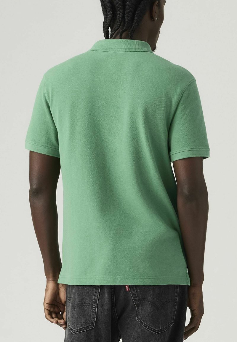 Hombre con cabello trenzado, vestido con una camiseta tipo polo verde de manga corta y jeans negros, de pie con la espalda hacia la cámara.