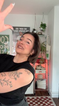 Svart ärmlös topp på en person med en detaljerad arm-tatuering. I bakgrunden syns en trådhylla med krukväxter och olika föremål.