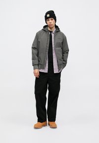 Γκρι τζάκετ με κουκούλα, φορεμένο πάνω από μια μαύρη μπλούζα και μια ριγέ πουκαμίσα. Μαύρο cargo παντελόνι και πορτοκαλί ξυλοπόδαρο συμπληρώνουν το ντύσιμο.