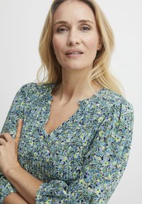 Blúzka s kvetinovým vzorom v modrej, zelenej a námorníckej farbe; má výstrih do V a zoskupené detaily. Dlhé rukávy s elastickými manžetami. Ľahká textúra materiálu.