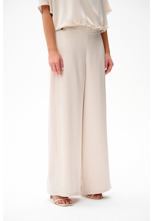 Kobieta ubrana w beżowe spodnie o szerokich nogawkach i dopasowane sandały, stojąca na białym tle.