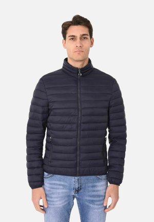 Giovane uomo con giacca trapuntata nera con cerniera e jeans azzurri, in piedi davanti a uno sfondo chiaro e uniforme.