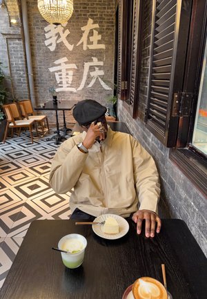 Άτομο που φοράει μπεζ μπουφάν και μαύρο καπέλο, καθισμένο σε τραπέζι καφετέριας με τσιζκέικ και δύο ποτά, μιλάει στο τηλέφωνο.