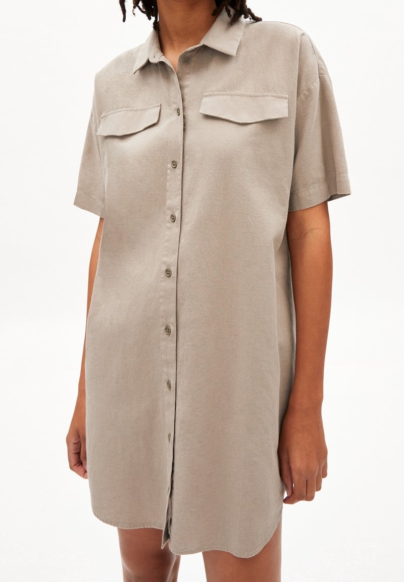 Robe chemise boutonnée en lin gris clair, avec des manches courtes, deux poches poitrine, et une coupe droite avec un ourlet arrondi.