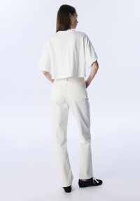 Weißes, kurzes T-Shirt und weiße High-Waist-Jeans. Glatter Stoff mit subtiler Textur. Gestylt mit schwarzen Schuhen, von hinten betrachtet.