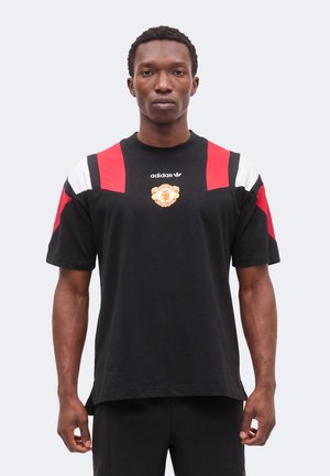 Man som bär en svart Adidas t-shirt med röda och vita axelpaneler och en Manchester United fotbollsklubb-logotyp på bröstet.