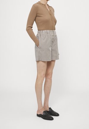 Ruskea pitkähihainen ribbineulospaita, jossa on napit, raidalliset korkeavyötäröiset shortsit ja mustat liukusukat. Yksinkertainen muotoilu, neutraali väripaletti.