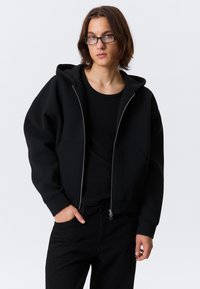 Felpa nera con zip realizzata in materiale morbido e con una vestibilità comoda. Presenta una tasca frontale e polsini a coste, indossata sopra una maglietta nera e pantaloni.