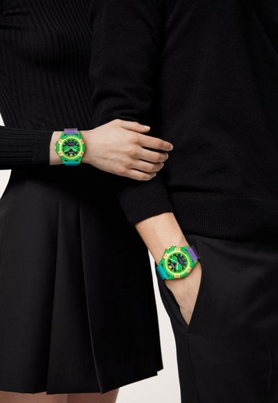 Dois relógios digitais com pulseiras verde e roxa apresentam um mostrador grande e preto com detalhes amarelos marcantes, usados nos pulsos de duas pessoas.