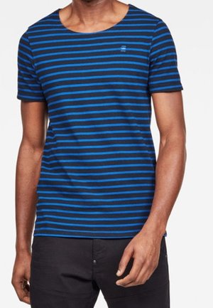Hombre que lleva una camiseta de manga corta con rayas horizontales azul marino y azul brillante, con pantalones negros, de pie con los brazos relajados.