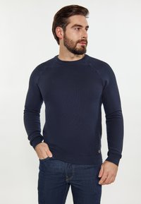 Pulover tricotat bleu marin cu guler rotund și sleeves raglan. Tesătură texturată și manșete ribbate, având un mic logo pe margine.