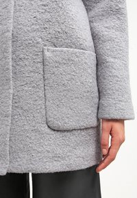 Manteau en laine gris clair texturé avec grande poche avant plaquée et manches longues, porté sur un pantalon noir, visible de mi-cuisse jusqu'en bas.