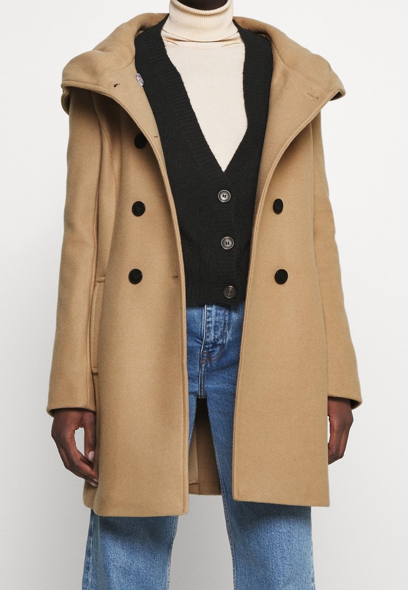 Cappotto in lana beige con un ampio cappuccio, design doppiopetto e bottoni neri. Indossato sopra un gilet di maglia nero e jeans blu.