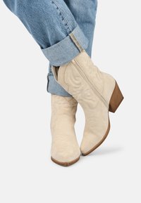 Botas de vaquero de gamuza beige con costuras decorativas, punta redonda y un tacón bajo de madera, llevadas con unos vaqueros de mezclilla azul cuffed.