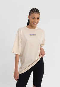 Beige oversized T-skjorte med korte ermer, med lilla grafisk tekst på brystet. Kombinert med svarte tettsittende shorts. Glatt stofftekstur.