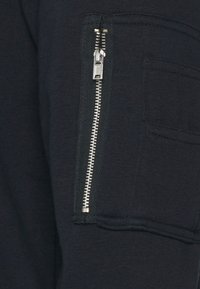 Tmavě modrá zipová kapsa na texturované tkanině, se stříbrným zipem a vedlejším detaily manžety, zdůrazňující funkční designové prvky.