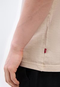 Κοντινό πλάνο ενός χεριού που ακουμπά δίπλα σε μια μπεζ πουκάμισο με κόκκινη ετικέτα Levi’s και μαύρο παντελόνι πάνω σε λευκό φόντο.