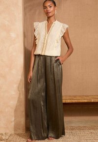 Cream-colored blouse with ruffled sleeves and button detail, paired with high-waisted, wide-leg, metallic bronze pants. Textured backdrop.
