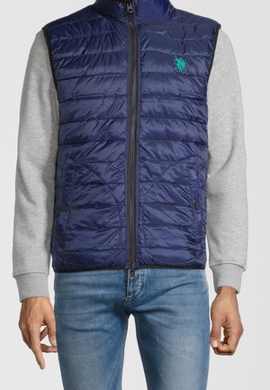 Homme portant un gilet matelassé bleu marine avec fermeture éclair à l'avant, sur une chemise grise à manches longues et un jean bleu, debout devant un fond uni.