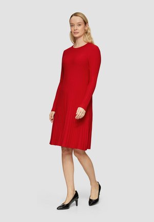Robe rouge à manches longues côtelées avec un col rond, une longueur mi-mollet et une silhouette ajustée, accompagnée de chaussures à talons hauts noires.