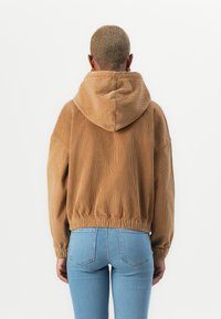Smeđi korduroj hoodie s uteznikom, elastičnom donjom ivicom i velikim džepom. Uparen s svijetloplavim uskim farmerkama. Glatka tekstura.