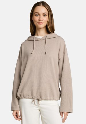 Jonge vrouw draagt een losse, lichtbeige hoodie met koordjes en een witte broek tegen een effen lichte achtergrond.