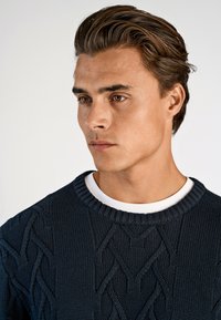 Marineblauer Strickpullover mit strukturiertem Zopfmuster, runder gerippter Ausschnitt, über einem weißen Hemd getragen. Glatte Stoffoberfläche.