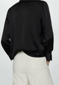 Blouse en satin noir avec un pli dans le dos, des poignets boutonnés et une coupe décontractée, associée à un pantalon taillé crème. Texture lisse, design minimal.