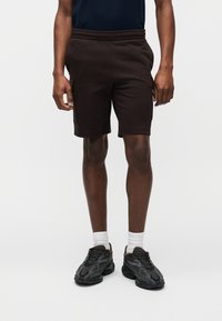 Άνδρας που φοράει μαύρα cargo σορτς, μαύρα χοντρά αθλητικά παπούτσια, άσπρες κάλτσες και σκούρο πουκάμισο σε απλό φωτεινό φόντο.