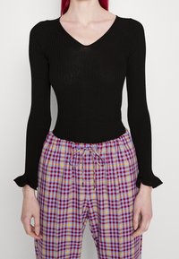 Kvinna klädd i en figurnära svart ribbad långärmad topp med volangärmar och flerfärgade röd, lila och gula rutiga byxor med snörning.