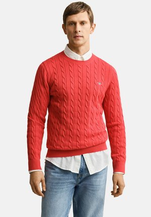 Uomo che indossa un maglione rosso a trecce sopra una camicia bianca con bottoni e jeans azzurri, in piedi davanti a uno sfondo semplice.