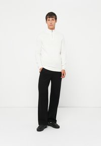 Weißer Zip-Up-Pullover mit geripptem Saum, kombiniert mit schwarzen weiten Hosen und schwarzen Schuhen. Das Modell steht vor einem schlichten weißen Hintergrund.