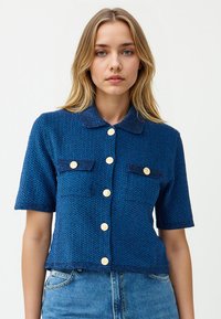 Junge Frau trägt ein blaues kurzärmeliges gestricktes Hemd mit goldenen Knöpfen und zwei Brusttaschen, kombiniert mit hellblauen Jeans.