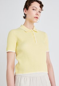 Polo a maniche corte giallo con texture a coste, colletto bianco e patta con bottoni. Design semplice senza motivi o accenti visibili.
