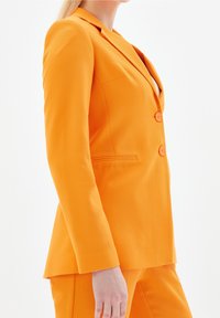 Pomarańczowa marynarka z fakturowanej tkaniny, z klapą z wycięciem, dwoma guzikami i pojedynczą kieszenią na piersi, pokazana z boku.