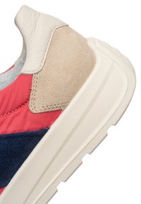 Nahaufnahme der Fersenseite eines Sneakers mit beigefarbenem Wildleder, weißem Leder, rotem Stoff, marineblauen Wildlederpaneelen und einer klobigen, cremeweißen Gummisohle.